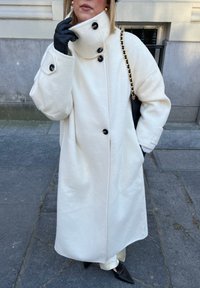 Femme en long manteau blanc avec de grands boutons noirs, gants noirs, chaussures pointues noires, et un sac à main noir avec une chaîne dorée sur un pavé en pierre.