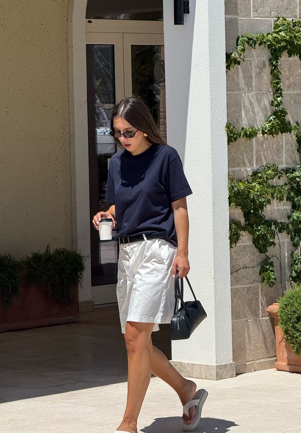 Femme portant des lunettes de soleil, une chemise bleu marine et un short blanc, marchant à l'extérieur en tenant une tasse de café et un sac à main noir près de l'entrée d'un bâtiment.