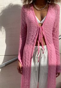 Rosa texturerad överdel med ett knyt i midjan, bärs över en vit bikini och kombineras med lätta, löst sittande vita byxor.