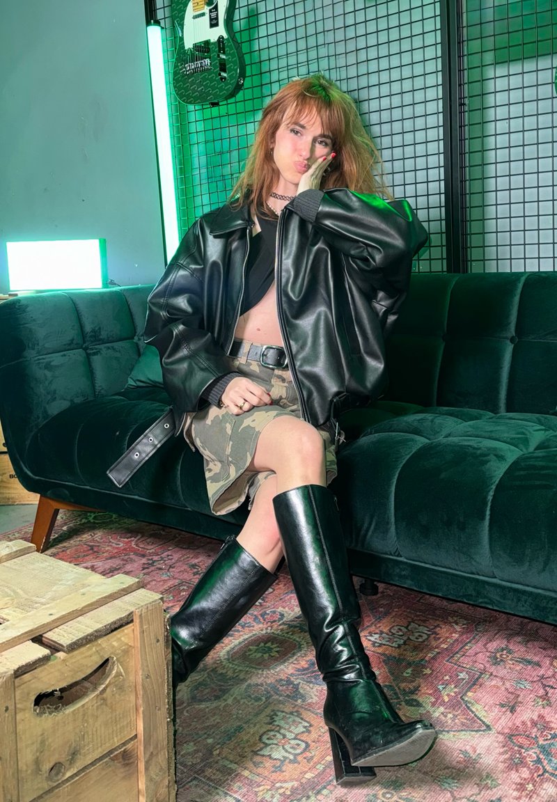 Giovane donna con giacca di pelle nera, gonna mimetica e stivali neri alti fino al ginocchio seduta su un divano verde con una chitarra appesa al muro.