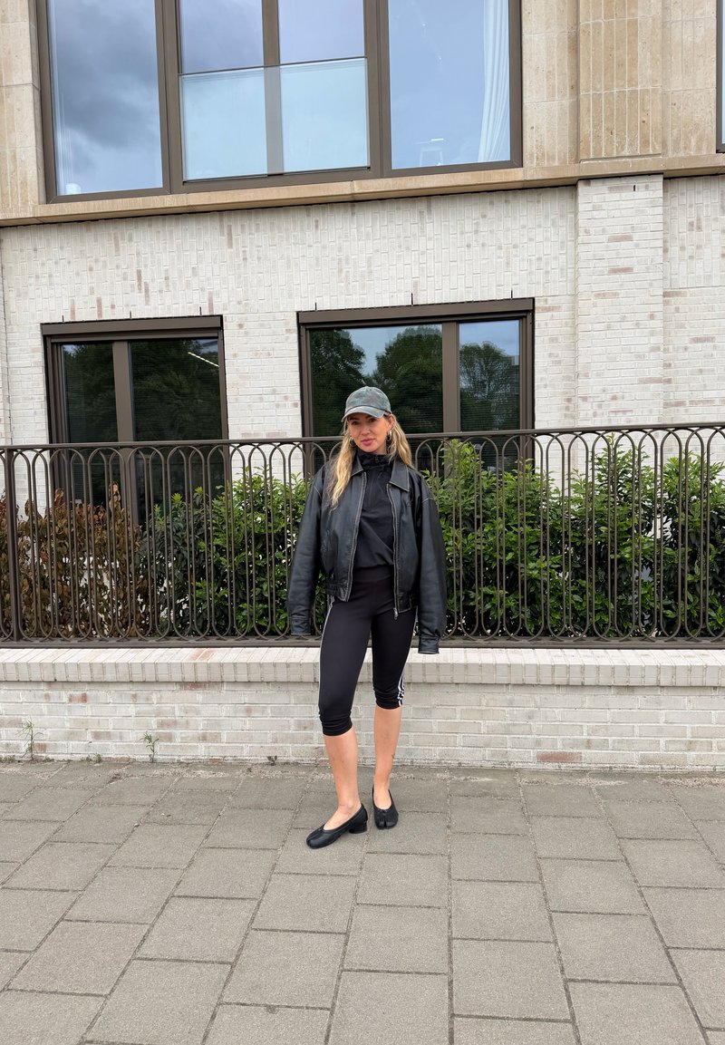 Junge Frau in schwarzen, gekürzten Leggings, Lederjacke, Kappe und flachen Schuhen steht am städtischen Gehsteig neben einem Geländer mit grünen Büschen und Fenstern im Hintergrund.