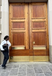 Kvinne med briller, hvit genser og svart veske står nær store trebredder dører med messingpaneler nederst.