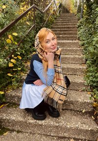 Écharpe beige à carreaux avec des franges, gilet bleu marine sur une chemise rayée bleu clair et une jupe blanche. Bottes noires imposantes, marches en pierre, verdure.