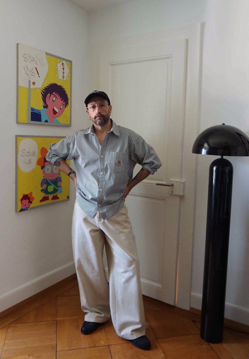 Homme en chemise rayée et pantalon beige large, debout les mains sur les hanches à côté d’une œuvre en style dessin animé « Save Us » et d’une lampe de sol noire moderne.