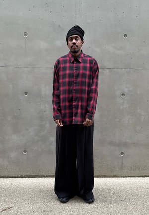 Hombre con pañuelo negro en la cabeza, camisa de cuadros roja y negra abotonada y pantalones anchos negros, de pie junto a una pared de hormigón gris.