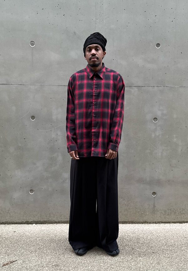 Homme portant un foulard noir sur la tête, une chemise à carreaux rouge et noire à boutons et un pantalon large noir, debout contre un mur en béton gris.