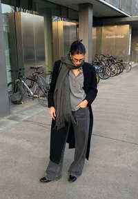 Juodas paltas, pilkai dryžuotas marškinėliai, pilka šalis ir pilki plačios kelnės. Nešiojami su juodais bateliais. Fone matomi dviračiai ir modernaus dizaino pastatas.