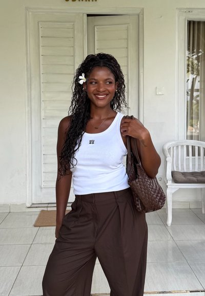 Mujer sonriente con cabello largo y rizado y flor blanca, vistiendo una camiseta sin mangas blanca y pantalones marrones, sosteniendo un bolso tejido marrón en interiores.