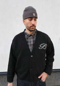 Cardigan noir et duveteux avec un col en V et quatre boutons, pr ésentant une lettre stylisée "B" grise sur le côté gauche, porté sur une chemise à carreaux.