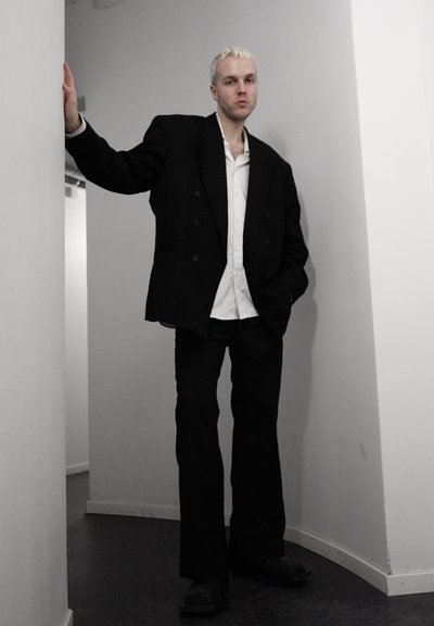 Joven con cabello rubio corto de pie en un pasillo blanco estrecho, vistiendo un blazer negro oversized, camisa blanca, pantalones negros y zapatos negros.