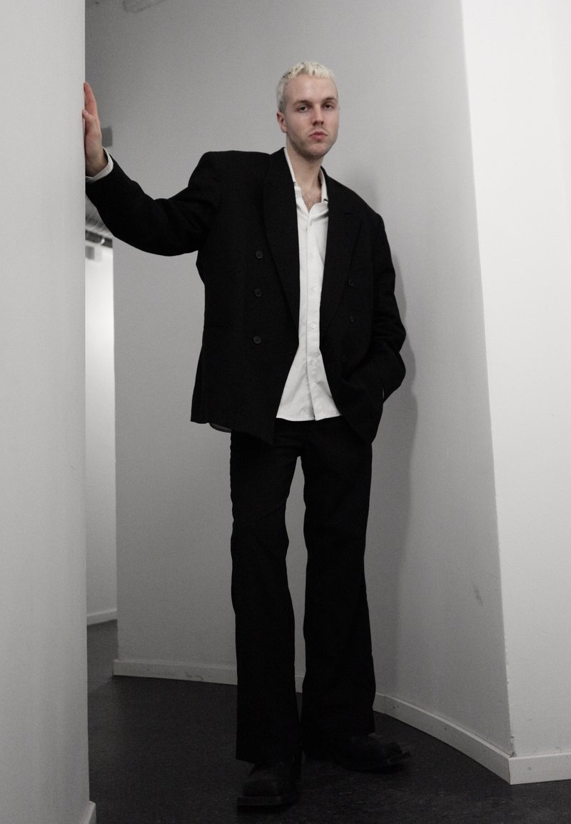 Jeune personne aux cheveux blonds courts, debout dans un couloir blanc étroit, portant un blazer noir surdimensionné, une chemise blanche, un pantalon noir et des chaussures noires.