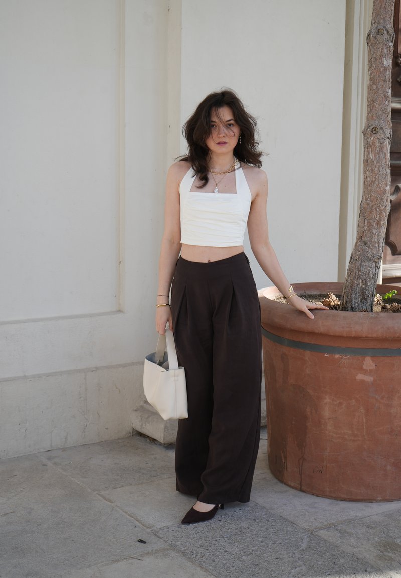 Mujer con cabello oscuro que lleva un top halter blanco recortado y pantalones anchos negros, sosteniendo una bolsa blanca, de pie junto a un gran árbol en maceta.