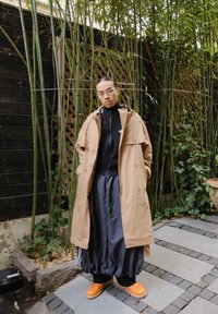 Trench-coat beige sur un pull en tricot noir, associé à un pantalon large noir et des sabots orange. Verdure texturée et chemin de pierre visibles.