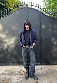 Crna zip-up hoodie, crna majica i sive traperice širokih nogavica. Crne sunčane naočale i crne čizme. Pozadina uključuje tamna kovana vrata.