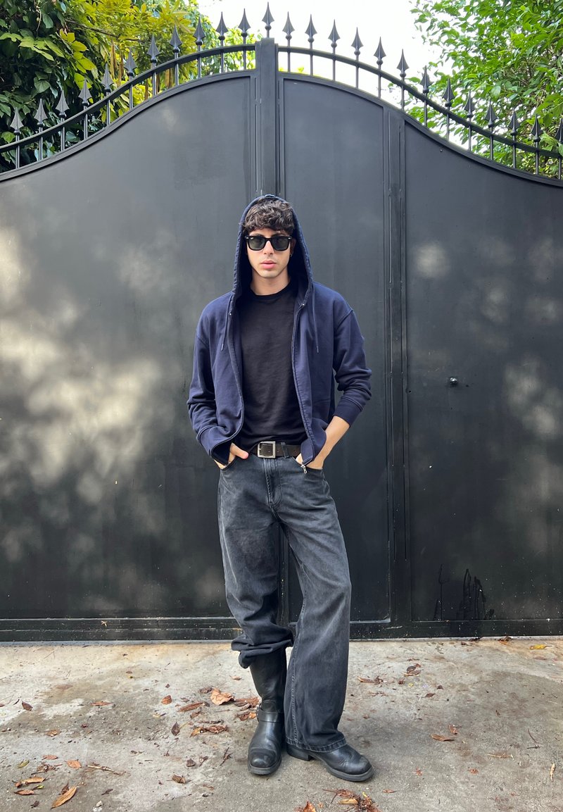 Zwarte zip-up hoodie, zwart t-shirt en grijze wijdvallende jeans. Zwarte zonnebril en zwarte laarzen. De achtergrond toont een donker smeedijzeren hek.