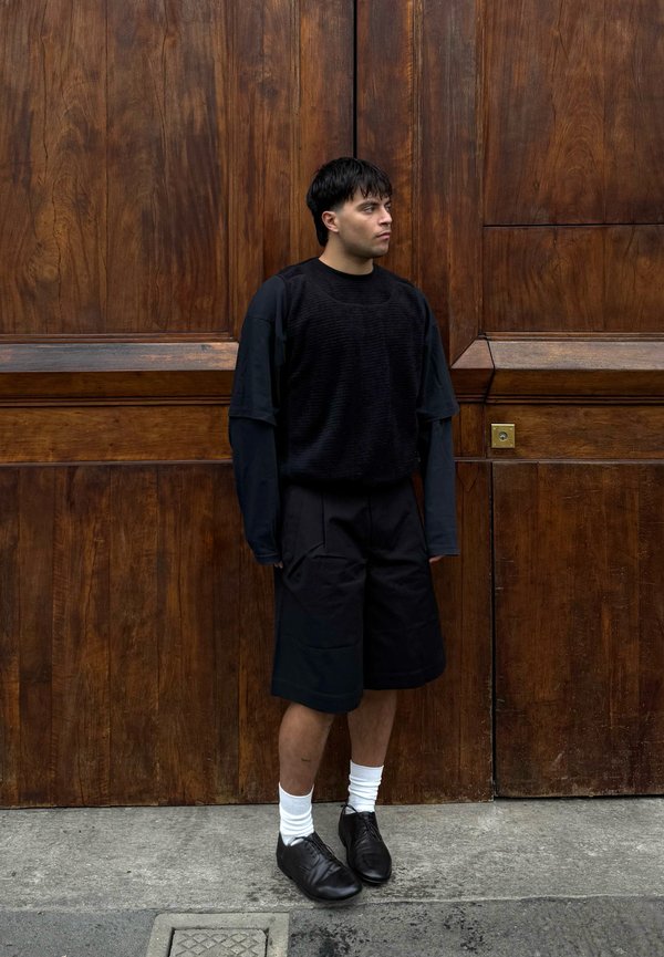 Young man in black layered outfit, white socks, and black shoes standing on concrete in front of large wooden doors, looking to the right.