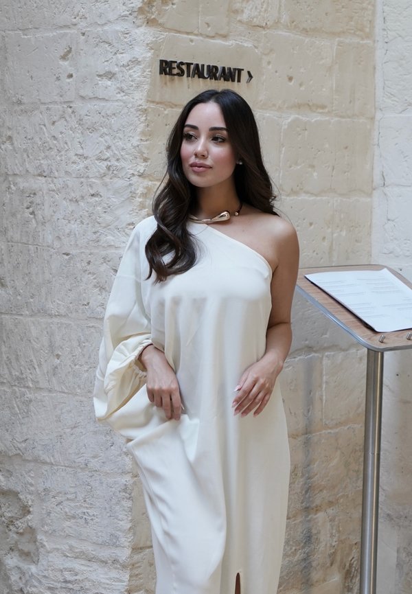 Donna con un vestito bianco monospalla in piedi vicino a un muro di pietra con un'insegna del ristorante e un menù su un espositore.