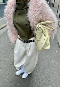 Personne portant une veste en peluche rose, un haut vert olive, un pantalon large blanc, des baskets blanches, une ceinture noire, tenant un sac à main jaune pâle dans une rue de la ville.