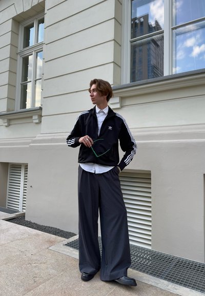 Chaqueta deportiva negra con cremallera y rayas blancas, camiseta blanca y pantalones grises anchos. De pie frente a un edificio de color claro.