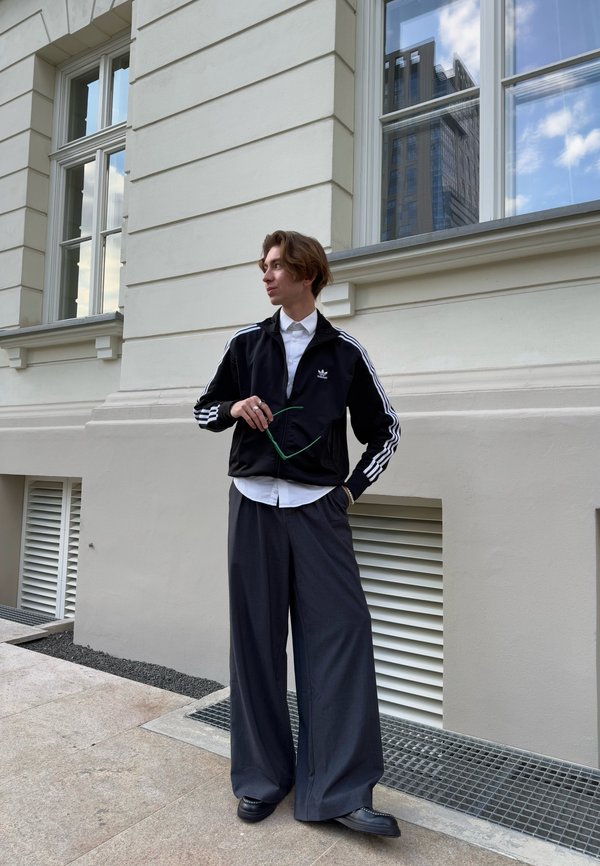 Črn športni zip-up jopič z belimi črtami, bela majica in široke sive hlače. Stoji pred svetlobnim zgradb.