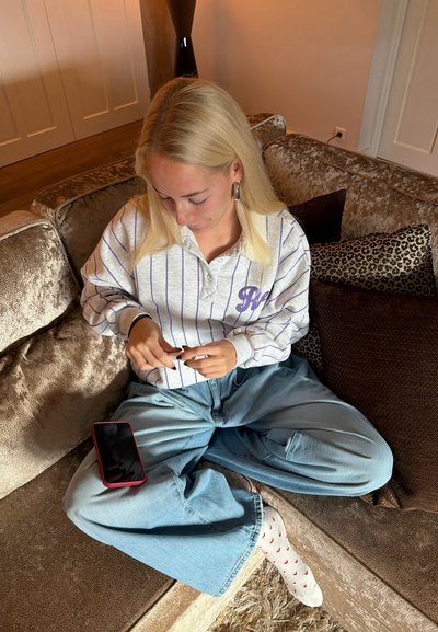 Una persona con cabello largo y rubio lleva una sudadera blanca con rayas y detalles morados, y unos vaqueros azules holgados, sentada en un sofá beige y mullido. Un teléfono inteligente está apoyado sobre su regazo.