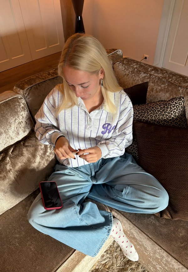 O persoană cu păr lung și blond poartă un hanorac alb cu dungi și accente mov și blugi largi albaștri, stând pe o canapea bej și moale. Un smartphone este așezat pe poala lor.