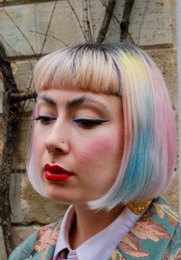 Kort pastelfarvet hår med farverige spidser, fremtrædende makeup med markant eyeliner og røde læber. Guld smykker fremhæver looket.