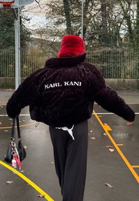 Personne portant un chapeau rouge et une veste noire Karl Kani, tenant un petit sac à main noir, marchant sur un terrain de basket en plein air.