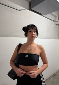 Mlada ženska z temnimi lasmi v bunih, oblečena v črn Adidas top in črne hlače, prilagaja svoj pas v enostavnem notranjem prostoru.
