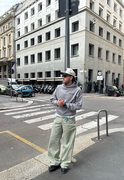 Sudadera gris, pantalones sueltos color verde claro y zapatos negros. Gorras de béisbol y gafas de sol. Escena urbana con edificios y vehículos.