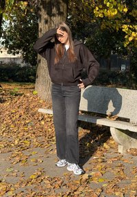 Sweat à capuche noir zippé, jean foncé et chaussures de sport blanches. Se tient sur un chemin couvert de feuilles tombées, à côté d'un banc en pierre sous un arbre.