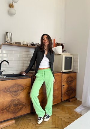 Mujer con chaqueta negra, camiseta blanca y pantalones verdes se apoya en la encimera de madera de la cocina, cerca del fregadero y una pequeña nevera.
