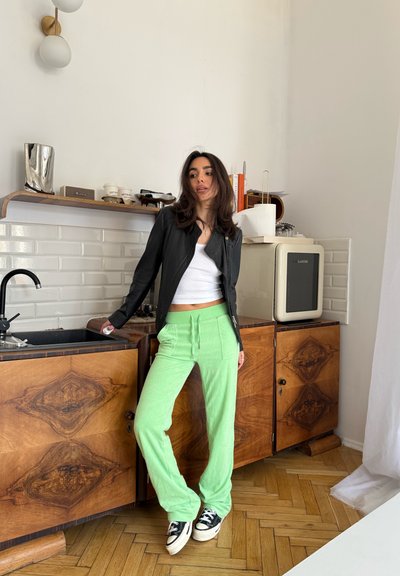 Mujer con chaqueta negra, camiseta blanca y pantalones verdes se apoya en la encimera de madera de la cocina, cerca del fregadero y una pequeña nevera.