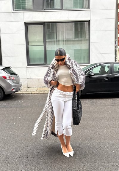Abrigo de piel sintética con rayas blancas y grises, top blanco ajustado, pantalones blancos cortos y tacones puntiagudos blancos. Bolso tejido negro y gafas de sol.
