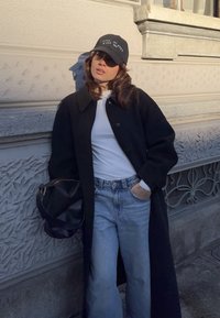 Γυναίκα που στηρίζεται σε υφασμένο τοίχο, φοράει μαύρο καπέλο, μαύρο πανωφόρι, λευκό πουκάμισο, μπλε τζιν παντελόνι, κρατώντας μαύρη τσάντα, με το ένα χέρι στην τσέπη.