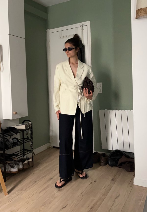 Femme portant un blazer crème oversize et un pantalon noir à jambes larges, tenant une pochette marron, debout à l'intérieur près d'une porte et d'un porte-chaussures.
