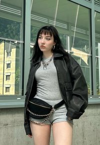 Nero giacca di pelle oversize, maglietta grigia attillata, pantaloni corti grigi e una borsa a tracolla nera con dettagli in catena. In basso si vedono grandi finestre.