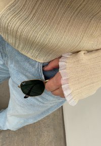 Χέρι σε τσέπη φωτεινού γαλάζιου τζιν που κρατάει γυαλιά ηλίου Ray-Ban με χρυσό πλαίσιο, φορώντας μπεζ πλεκτό πουλόβερ με μανσέτες με βολάν.