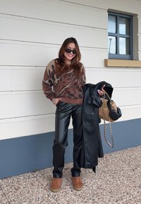 Καφέ και μπεζ πουλόβερ με σχέδιο, μαύρο δερμάτινο παντελόνι, καφέ υποδήματα, κρατώντας ένα μαύρο παλτό και μια καφέ τσάντα με χρυσή αλυσίδα.