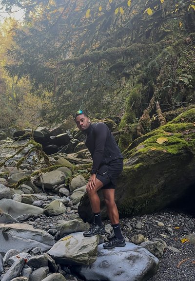 Persona con una camiseta deportiva negra de manga larga y pantalones cortos, de pie sobre grandes rocas grises, rodeada de rocas cubiertas de musgo y follaje verde.