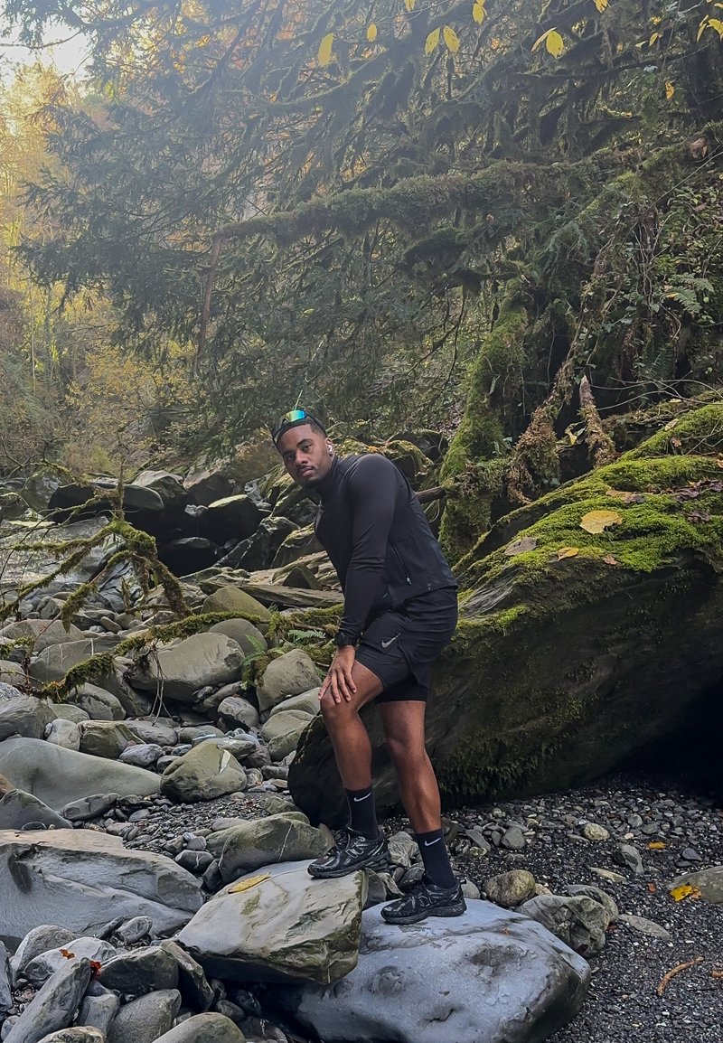 Una persona con una maglietta nera sportiva a maniche lunghe e pantaloncini è in piedi su grandi rocce grigie, circondata da rocce coperte di muschio e vegetazione verde.