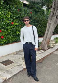 Hvid langærmet skjorte, marineblå stribede vide bukser, sorte lædersko. Stående mod en grøn bladbaggrund med røde blomster.