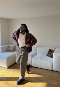 Homme souriant portant une doudoune bordeaux, un t-shirt blanc, un pantalon gris et des bottes noires, debout dans un salon lumineux avec un canapé blanc.