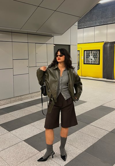 Mujer con gafas de sol, chaqueta de cuero, top gris, pantalones cortos negros y tacones con calcetines, de pie en un metro cerca de un tren amarillo con un símbolo de silla de ruedas.