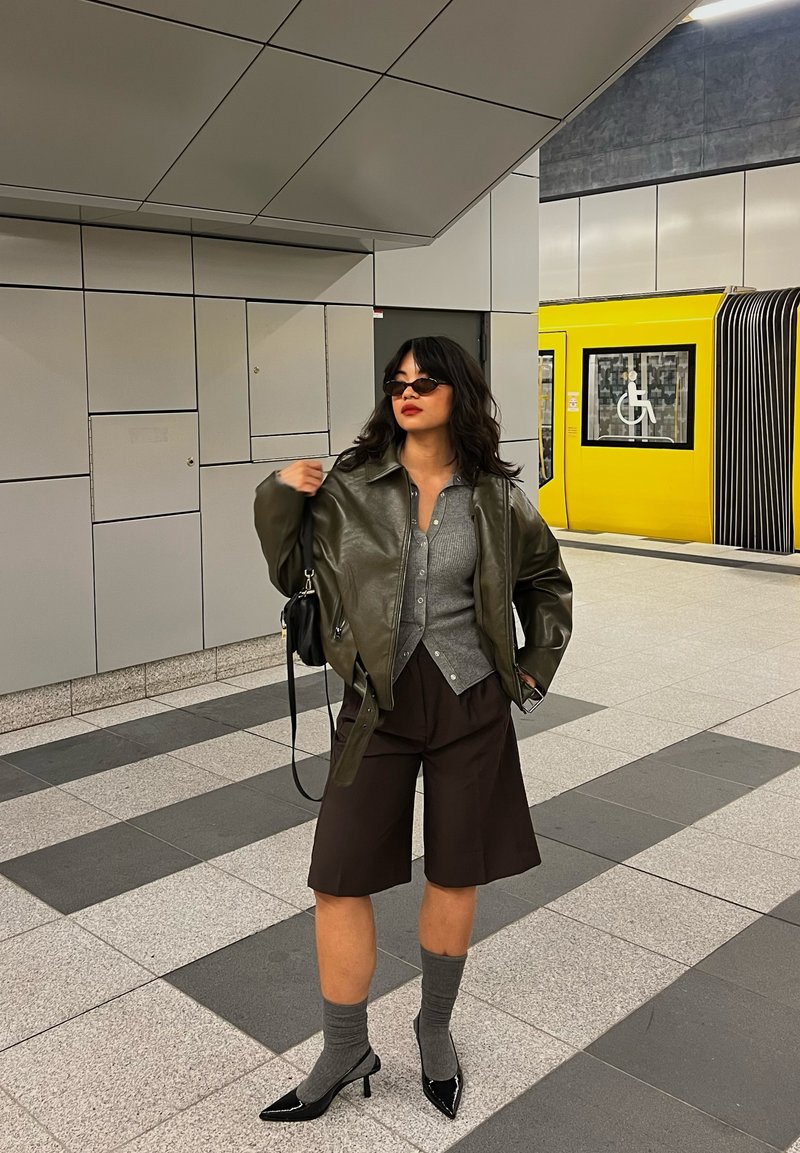 Mulher usando óculos de sol, jaqueta de couro, blusa cinza, calções pretos e saltos com meias, de pé numa estação de metro perto de um comboio amarelo com um símbolo de cadeira de rodas.