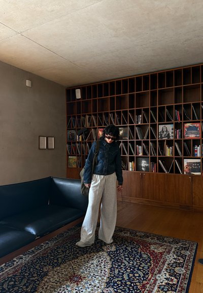 Chaqueta de mezclilla, pantalones de chándal grises de pierna ancha, sofá negro sobre una alfombra estampada. Estantes de madera con libros y discos en un espacio interior minimalista.