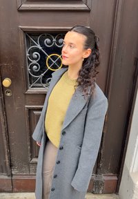 Manteau en laine gris avec un grand col et des boutons, superposé à un pull en maille épaisse vert olive, associé à une jupe beige élégante.