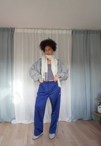 Sudadera gris de gran tamaño, pantalones deportivos azules con rayas blancas, bufanda crema y zapatos grises. Texturas suaves y ajuste relajado.