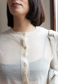 Ανοιχτό μπεζ ζακέτα με στρογγυλή λαιμόκοψη και σχέδιο με κουμπιά μπροστά. Διαθέτει υφή πλεκτού, συνδυασμένη με λευκή τσάντα με χερούλι στον ώμο.