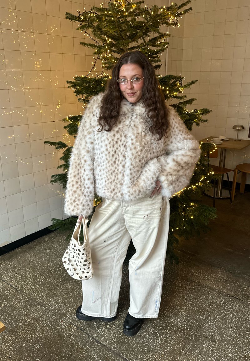 Frau mit langen lockigen Haaren, die eine gepunktete Kunstpelzjacke und weiße Hosen trägt, steht drinnen und hält eine weiße Handtasche neben einem geschmückten Weihnachtsbaum.